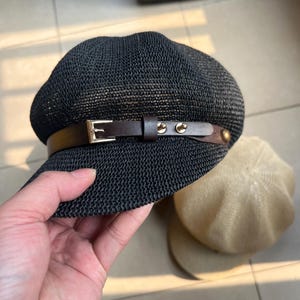 May include: Black newsboy cap with a woven texture, featuring a brown leather band with a gold buckle and rivets. A tan beret is partially visible in the background. The cap is held by a hand.