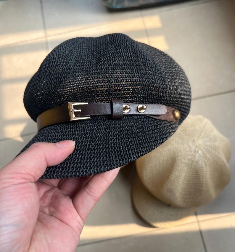 May include: Black newsboy cap with a woven texture, featuring a brown leather band with a gold buckle and rivets. A tan beret is partially visible in the background. The cap is held by a hand.