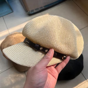 May include: A beige newsboy cap with a woven texture and a dark brown leather band. The band is adorned with gold-colored rivets and a buckle. The cap has a structured brim and a rounded crown, offering a stylish accessory.
