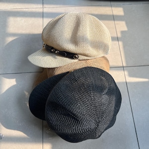 May include: Three hats in a stack. The top hat is beige with a black band and silver studs. The middle hat is tan, and the bottom hat is black. All hats have a woven texture.