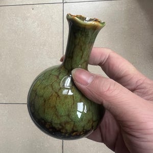 Puede incluir: Un pequeño jarrón redondo de cerámica verde con cuello estrecho y borde acampanado. El jarrón tiene un aspecto moteado y texturizado con acentos marrones. El jarrón se sostiene en una mano, mostrando su tamaño y forma. El jarrón es probablemente para fines decorativos.