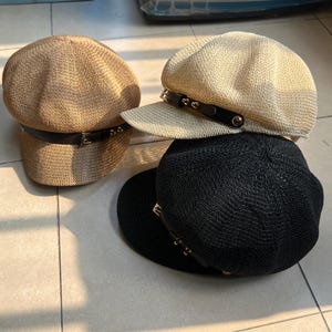 May include: Four woven newsboy caps in tan, cream, and black. Each cap features a black leather band with a metal buckle and decorative studs. The caps are stacked on a tiled surface, showcasing their different colors and styles.