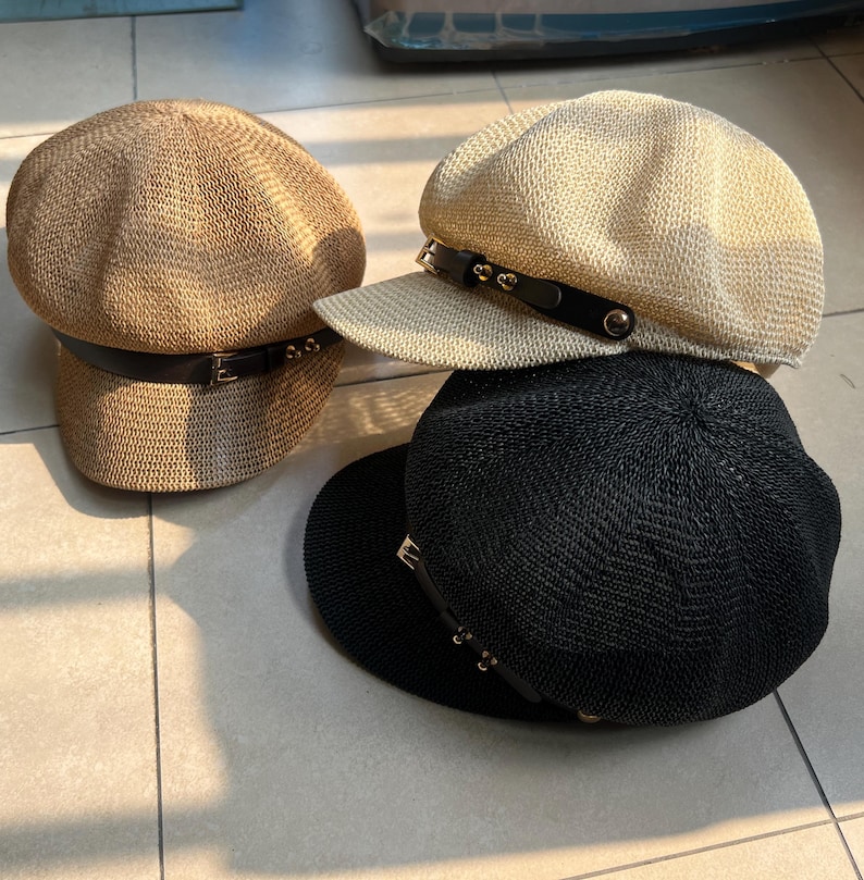 May include: Four woven newsboy caps in tan, cream, and black. Each cap features a black leather band with a metal buckle and decorative studs. The caps are stacked on a tiled surface, showcasing their different colors and styles.