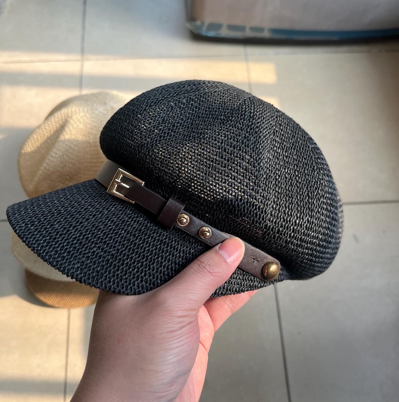 May include: A dark navy blue woven hat with a brown leather band and gold-colored rivets. The hat has a short brim and a rounded crown. A second, light-colored hat is partially visible in the background.