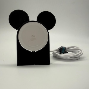 May include: Black wireless charging stand in the shape of Mickey Mouse ears. The stand has a white charging pad and a white cord. The cord is secured with a green and black cable tie.