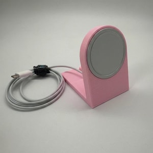 May include: A pink charging stand with a silver charging pad. A white charging cable is connected to the stand and coiled next to it. The stand is designed to hold a charging device.