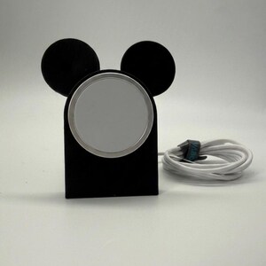 May include: Black wireless charger in the shape of Mickey Mouse ears. The charger has a silver charging surface and comes with a white charging cable. The design is a fun and functional accessory for any desk or nightstand.