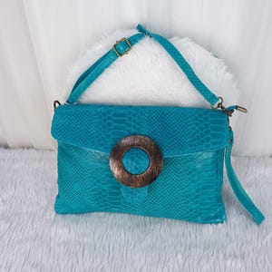 Op de afbeelding: Een turquoise clutch met een slangenpatroon en een grote, ronde houten gesp. De tas heeft een bijpassende schouderriem en een klepsluiting. De achtergrond is een wit, pluizig oppervlak.