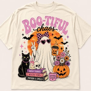 May include: A cream-coloured t-shirt featuring a graphic of a ghost wearing a chequered bow and heart-shaped sunglasses, holding a coffee cup and making a peace sign. The design includes a black cat, a skull, books, a pumpkin, and the text BOO-TIFUL chaos.