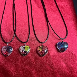 May include: Four heart-shaped pendant necklaces with black cords. Each pendant features a colorful tree design encased in clear resin. The trees display shades of red, green, yellow, and blue. The necklaces rest on a red fabric surface.