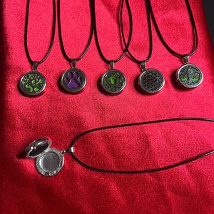 May include: A collection of pendant necklaces with black cords, featuring various designs within silver-toned lockets. Designs include a green tree, purple paw print, green lotus, geometric pattern, and a tree of life. One open locket is also shown.