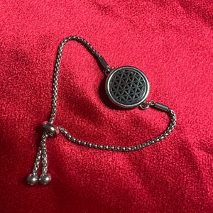 May include: A silver-toned bracelet with a circular pendant featuring a black flower of life design. The adjustable chain bracelet has a sliding bead closure and hangs on a red surface.