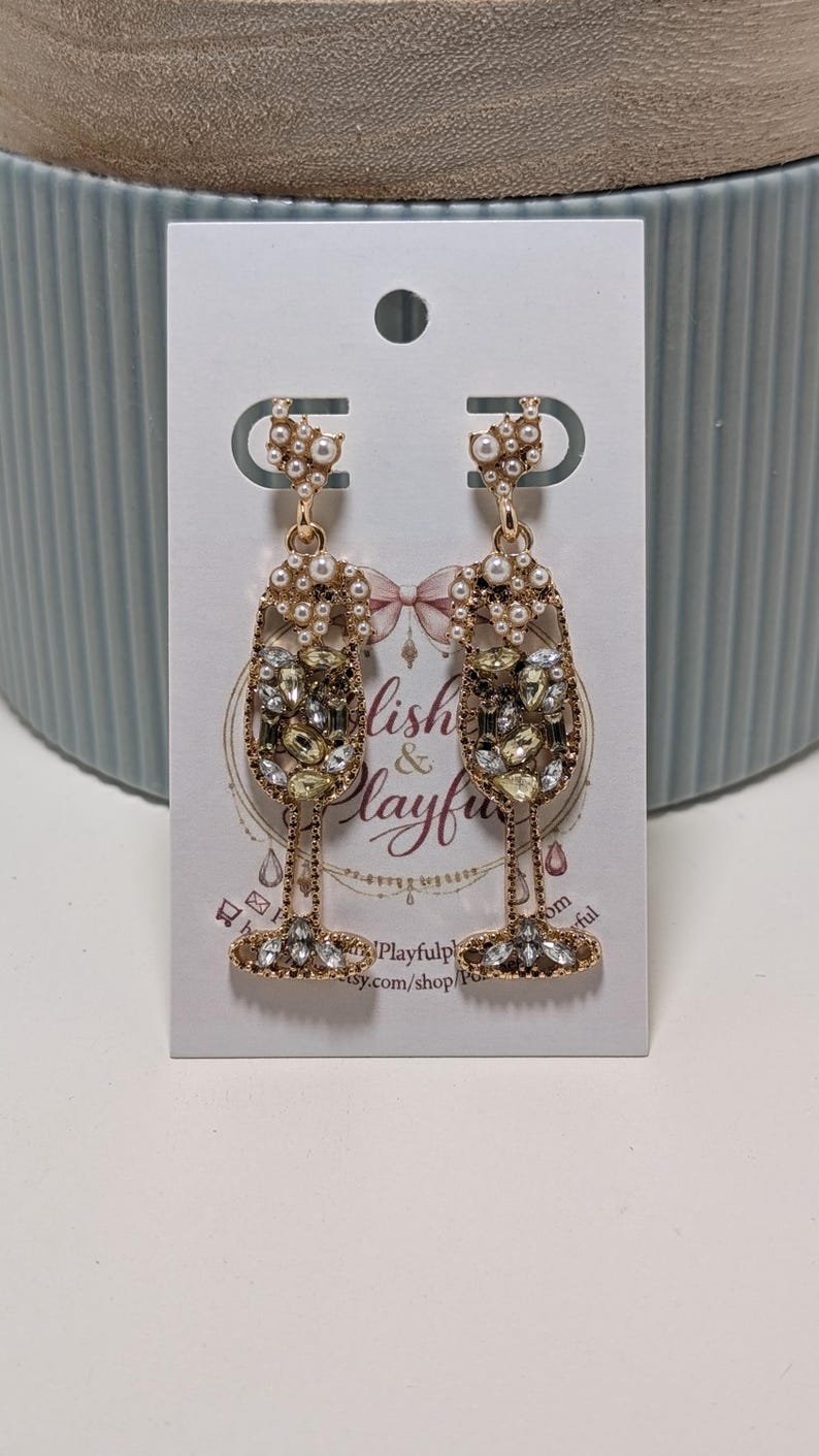 Puede incluir: Un par de pendientes con forma de copa de champ&aacute;n doradas, adornados con cristales transparentes y color champ&aacute;n, y perlas de imitaci&oacute;n. Los pendientes se exhiben en una tarjeta blanca con el texto "Lish & Playful".