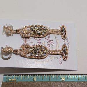 Puede incluir: Un par de pendientes con forma de copa de champ&aacute;n. Los pendientes son dorados e incrustados con pedrer&iacute;a transparente y color perla. Los pendientes se exhiben en una tarjeta blanca con texto y un dise&ntilde;o floral.