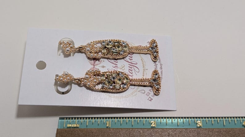 Puede incluir: Un par de pendientes con forma de copa de champ&aacute;n. Los pendientes son dorados e incrustados con pedrer&iacute;a transparente y color perla. Los pendientes se exhiben en una tarjeta blanca con texto y un dise&ntilde;o floral.