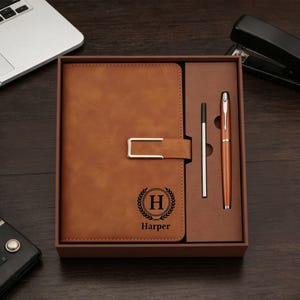 May include: A brown leather journal and pen set in a gift box. The journal has a silver clasp and a monogram with the letter "H" and the name "Harper". A silver pen and a brown pen are included.