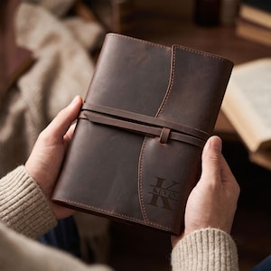 May include: A dark brown leather journal featuring a wrap-around leather strap closure and a vertical flap design. The front cover is engraved with a large letter K and the name KEVIN centered horizontally across the letter in a serif font.