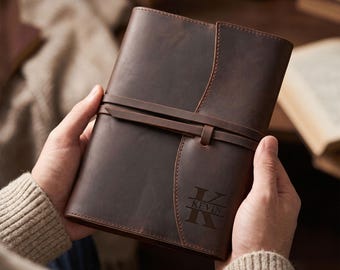 May include: A dark brown leather journal featuring a wrap-around strap closure and a vertical flap design. The front cover is personalized with a large letter K and the name KEVIN engraved in a serif font. The leather has a smooth, matte finish.