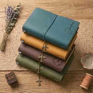 Op de afbeelding: Een stapel leren tijdschriften in teal, mosterd, bruin en olijfgroen. Het teal tijdschrift heeft de naam "Jordan" in reliëf op de omslag. Elk tijdschrift heeft een bijpassende elastische sluiting en een bedel van kleine vliegtuigen. Een bosje lavendel en een houten spoel zijn ook zichtbaar.