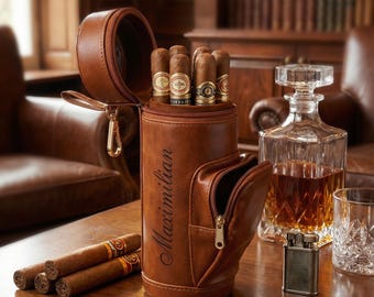 May include: A brown leather cigar case with the name "Maximilian" inscribed on the side, holding several cigars. A matching pocket with a zipper is on the front. Several cigars are on the table, along with a glass decanter, a lighter, and a glass.
