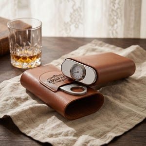 May include: A brown leather cigar case with a silver-toned metal end cap featuring an integrated analog hygrometer. The case includes a silver-toned metal cigar cutter stored in a front pocket, which is embossed with the name ALEXANDER.