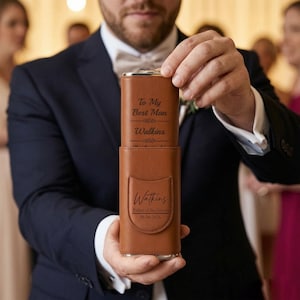 May include: A brown leather-like flask case with a flap, engraved with the text 'Watkins', 'Father of the Groom', and '08-06-2026'. A matching brown flask is partially pulled out, showing the engraved text 'To My Best Man' and 'Watkins'.