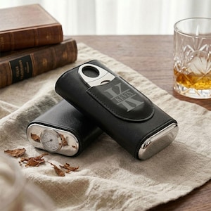 May include: A black leather cigar case featuring a silver-toned metal cigar cutter and a built-in analog hygrometer. The case is engraved with the name KEVIN in a bold, stylized font. The design includes a flap closure and polished metal ends.