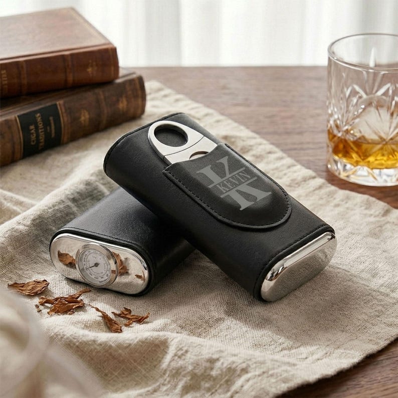 May include: A black leather cigar case featuring a silver-toned metal cigar cutter and a built-in analog hygrometer. The case is engraved with the name KEVIN in a bold, stylized font. The design includes a flap closure and polished metal ends.