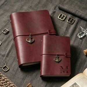 May include: Two burgundy leather journals with anchor charms and elastic closures. The larger journal has a vertical orientation, while the smaller one has personalized text. The journals are on a gray surface.