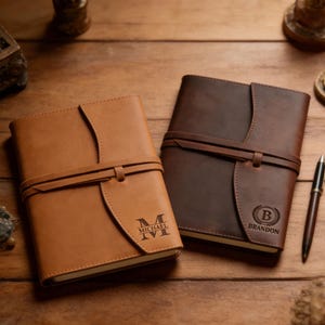 May include: Two leather journals, one tan and one dark brown, lie on a wooden surface. The tan journal has the name "MICHAEL" and the letter "M" engraved. The dark brown journal has the name "BRANDON" and the letter "B" engraved. Both journals have a strap closure.