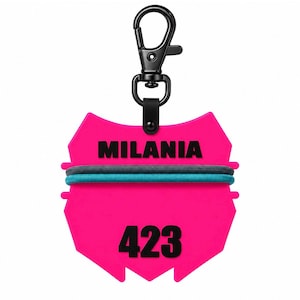 May include: A bright pink, shield-shaped plastic tag featuring the name MILANIA and the number 423 in bold black text. Two elastic hair ties, one dark gray and one teal, are wrapped around the center. A black metal lobster clasp is attached.