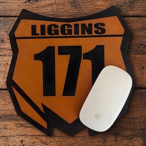 以下が含まれることがあります： 「LIGGINS」の文字と数字「171」が黒で描かれた、茶色と黒の盾型のデスクマット。Appleのロゴが入った白いコンピューターマウスがマットの隣にあります。背景は木製の表面です。