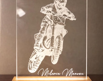 May include: A rectangular clear acrylic plaque featuring an etched graphic of a motocross rider on a dirt bike, mounted on a rectangular light-colored wooden base. The name Milania Monroe is etched in a cursive script at the bottom of the plaque.