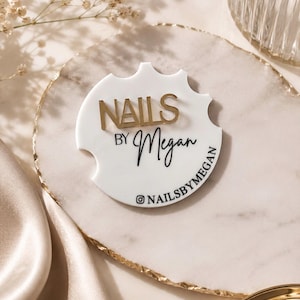 May include: A circular white acrylic sign with a notched edge featuring the gold raised text NAILS BY and the black script text Megan, with the Instagram handle @NAILSBYMEGAN printed in black along the bottom curve of the sign.