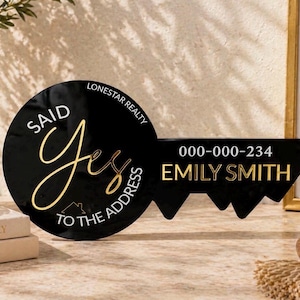 May include: A black, key-shaped sign featuring gold text that reads "SAID Yes TO THE ADDRESS", "LONESTAR REALTY", "000-000-234", and "EMILY SMITH". The sign includes a small gold house icon and is designed for real estate closing photos.