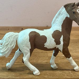 May include: A painted toy horse figurine in a walking pose. The horse has a brown body with white patches and a white mane and tail. The horse is standing on a wooden surface.