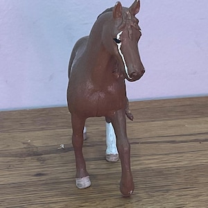 May include: A brown toy horse figurine with white markings on its face and legs. The horse is standing on a wooden surface, with a light purple wall in the background. The horse is a detailed miniature.