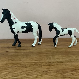 May include: Two toy horses, a mare and foal, are depicted. The mare is black and white with a black mane and tail, and the foal has a similar pattern. The mare has red accents in her mane. The toys are on a wooden surface.