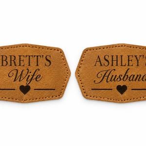 May include: Two tan, hexagonal-shaped patches with stitched edges. The left patch features the text "BRETT'S Wife" above a small heart icon. The right patch features the text "ASHLEY'S Husband" above a small heart icon. Both have dark brown lettering.
