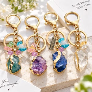 May include: Four gold-toned metal keychains featuring raw, electroplated crystal pendants in green, purple, blue, and clear. Each keychain includes a lobster clasp, a circular ring, and small colorful gemstone beads. One keychain has a rectangular tag.
