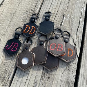 May include: A collection of personalized keychains with black, brown, and tan leather tags. Each keychain features embroidered initials in various colors, including orange, pink, and white, attached to a metal ring.