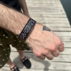 Pode incluir: Pulseira de couro preta com a data "08/03/25" bordada em fio rosa. A pulseira é usada em um pulso com tatuagens visíveis. O fundo mostra um cais de madeira e água.