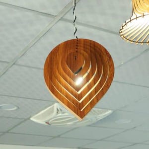 Puede incluir: Una lámpara colgante de madera con un diseño en forma de hoja. La lámpara está hecha de madera en capas, creando un brillo cálido y acogedor. La luz cuelga de un cable blanco y negro y está suspendida de un techo blanco. Otra lámpara es visible en el fondo.