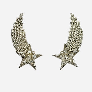 May include: A pair of silver-toned star earrings. Each earring features a five-pointed star with clear crystal accents and a curved, cascading design of smaller crystals, creating a shooting star effect. The earrings are designed for pierced ears.