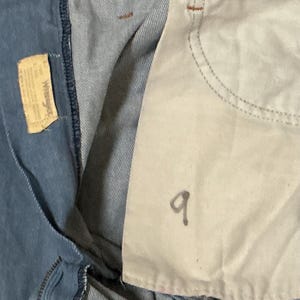 May include: Close-up of denim fabric with a light blue hue. A small, rectangular tag with text is visible on the left. The number "9" is written in black ink on the right side of the image.