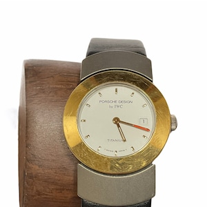 May include: A gold and silver wristwatch with a white face and black leather strap. The watch face says "Porsche Design by IWC" and "Titanium".