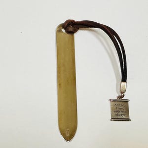 Op de afbeelding: Een goudkleurige metalen bladwijzer met een bruin leren koord en een kleine rechthoekige bedel. De bedel is gegraveerd met de woorden "And lo, I am, with you always."