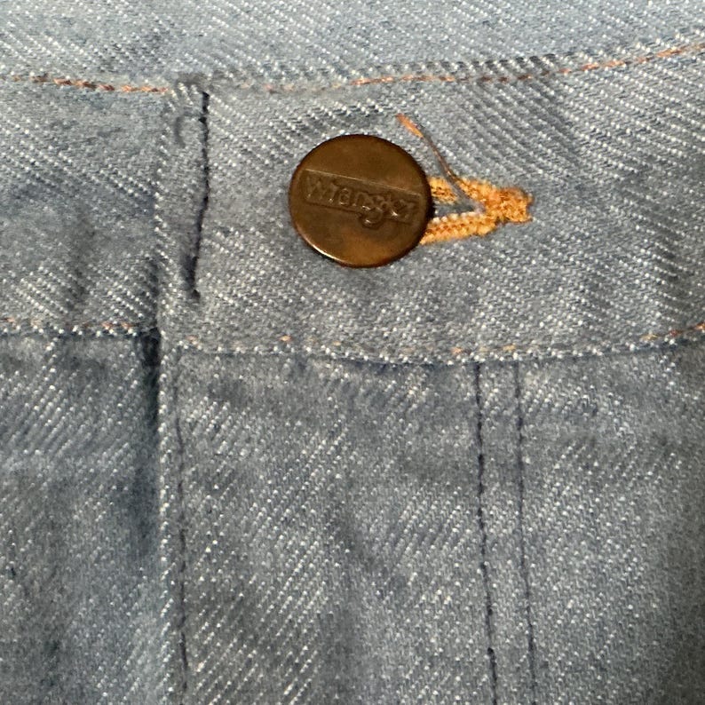 May include: Close-up of light blue denim fabric with a brown button. The button is round and has the word "Wrangler" embossed on it. The denim has visible stitching and a classic texture.