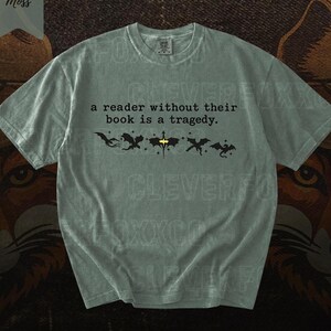May include: A sage green t-shirt with the text "a reader without their book is a tragedy." Below the text are black dragon silhouettes. The shirt has a crew neck and short sleeves. The word "Moss" is in the upper left corner.