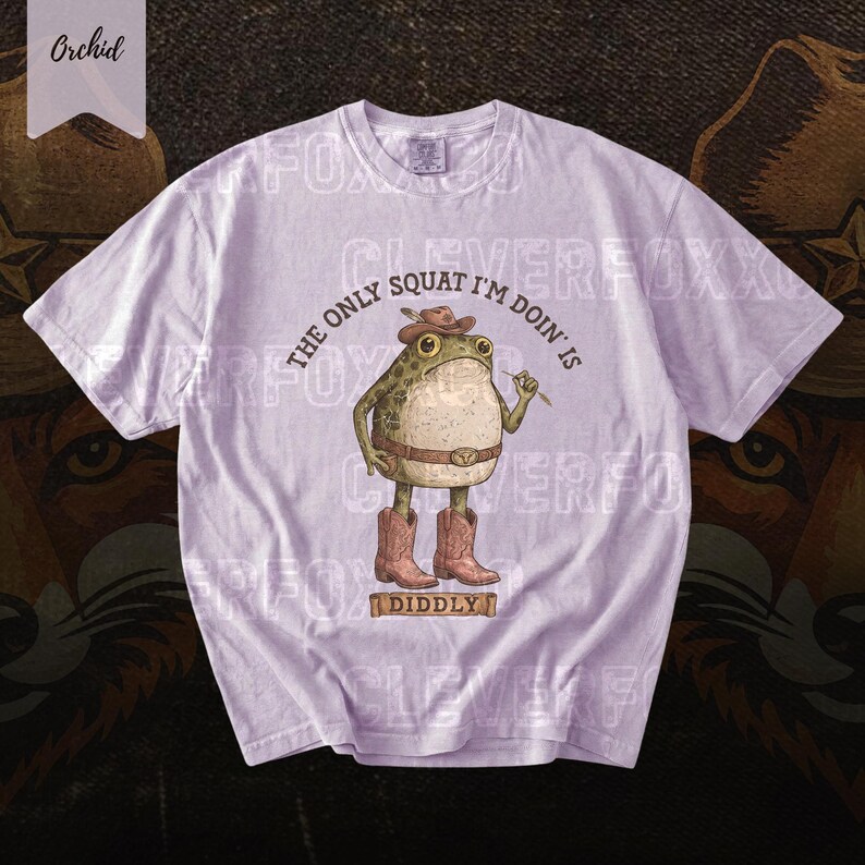 A white t-shirt with a cartoon frog wearing a cowboy hat and boots, along with the text "The only squat I'm doing is Diddlyy" printed on it.